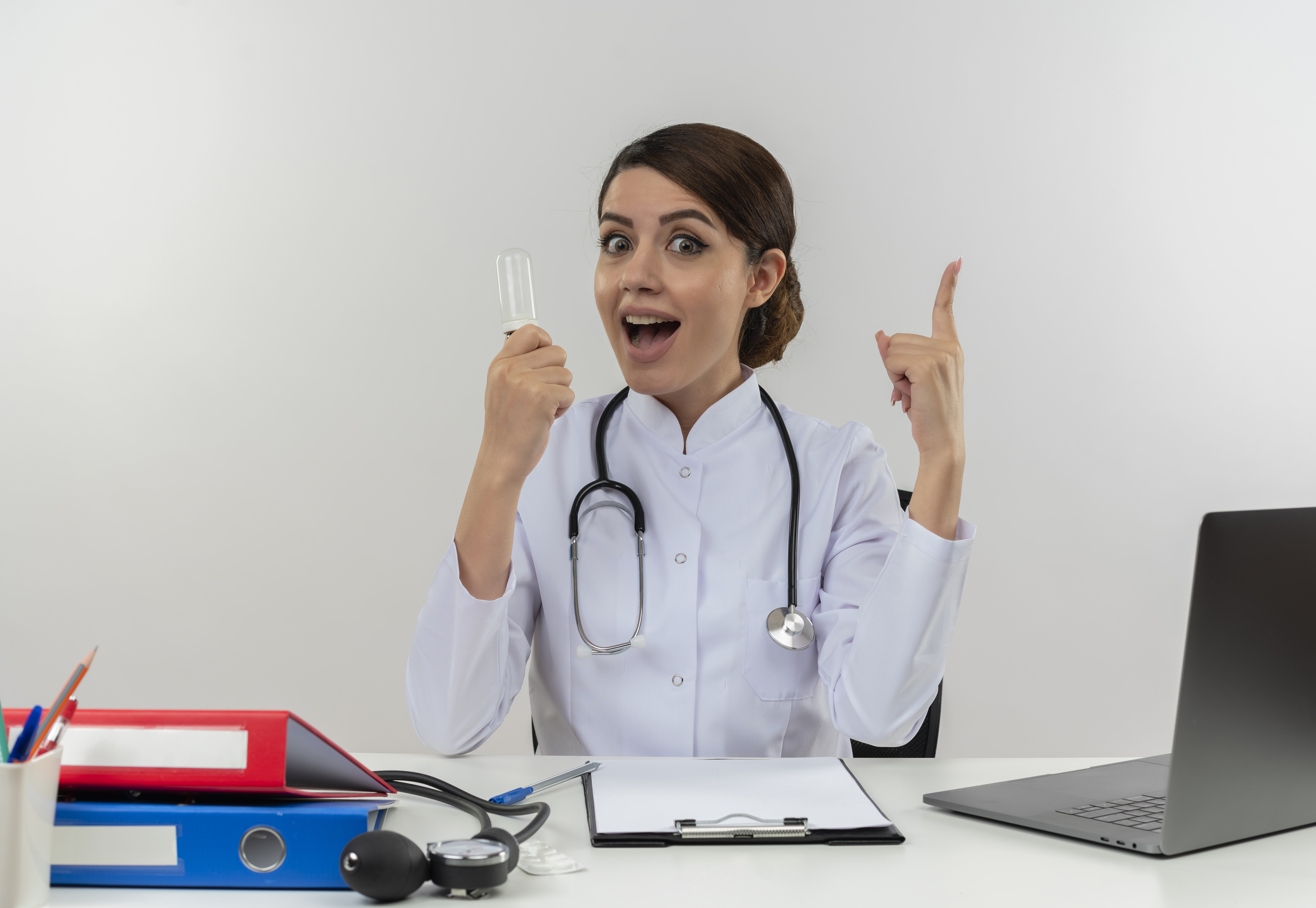 joyful young female doctor wearing medical robe with stethoscope sitting at desk work on computer with medical tools holding light bulb her thumb up on isolated white background with copy space